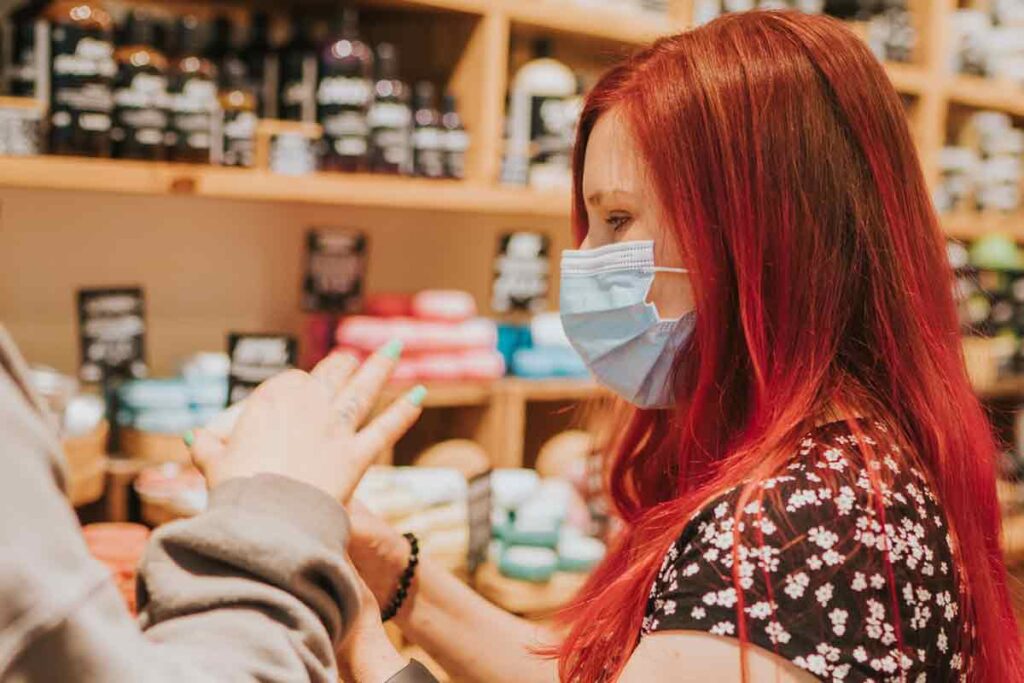 Sassy having long red hair, wearing a mask, and a black dress with small flowers. She is in a shop, and the shop assistant is helping her choose a product. You can only see the hands of the shop assistant. The product is in both their hands, and can't be identified.