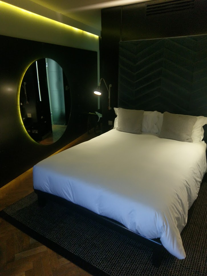 A large Bed with white sheets, a large circular mirror is hanging on the wall in the left of the picture.
