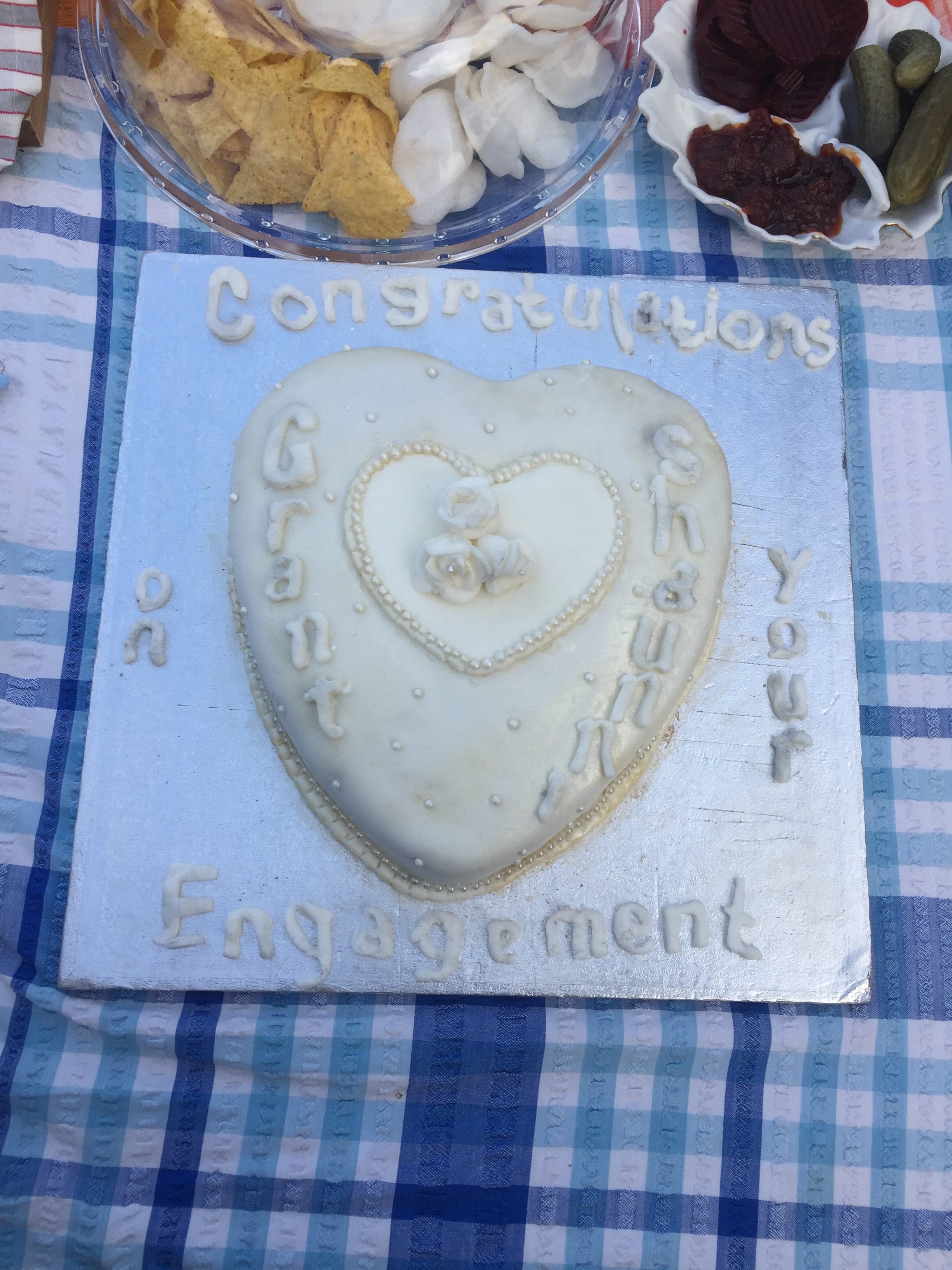 A white iced Heart Shaped cake. There is another smaller heart in the middle of the cake and some roses in the center of that. The cake reads congratulation on your engagement. The coupls name are on either side of the heart in the middle.
