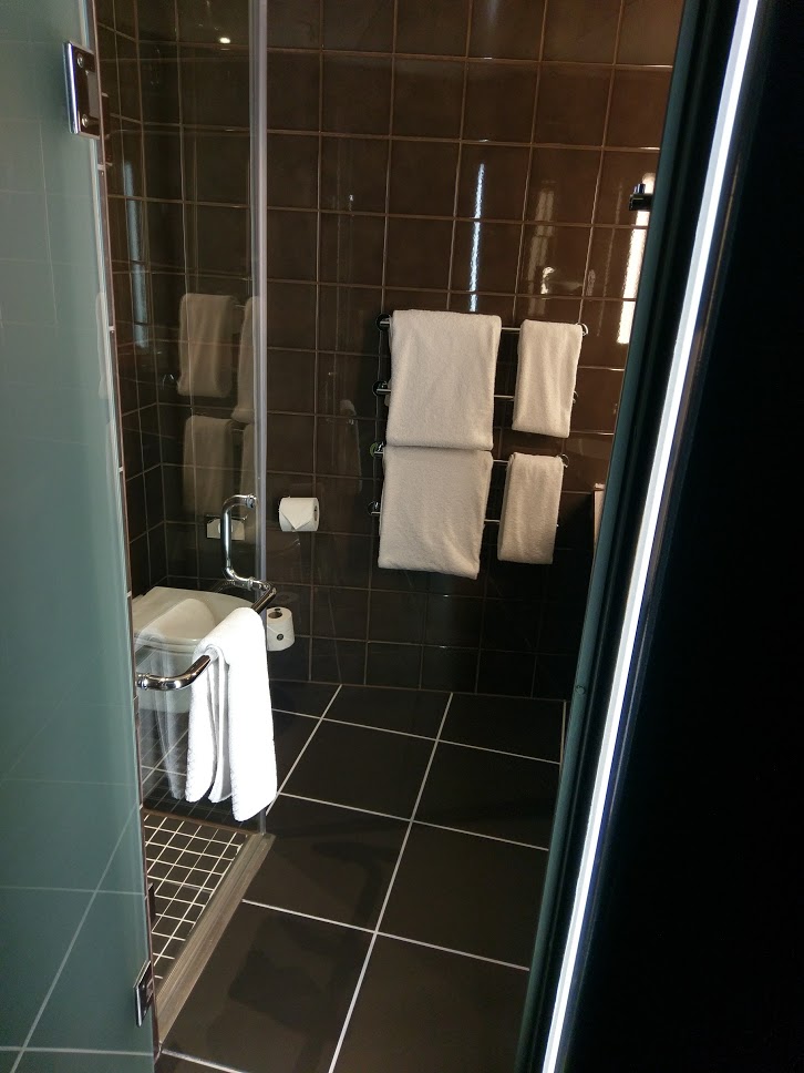 The bathroom in the hoxton Shoreditch hotel room. There is a walk-in rainfall shower on the left and the sink is on the right, black tiles cover the floor and walls