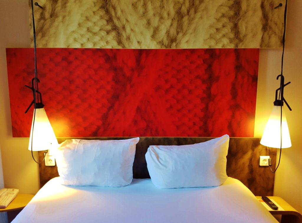 A double bed with white sheets and a big red headboard. There are two bedside tables one at each side and two lamps hanging down about each table.