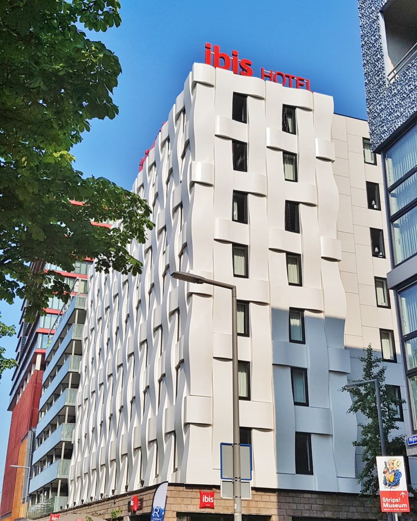 A 7 story white building with big red letters spelling out Ibis Hotel on the top