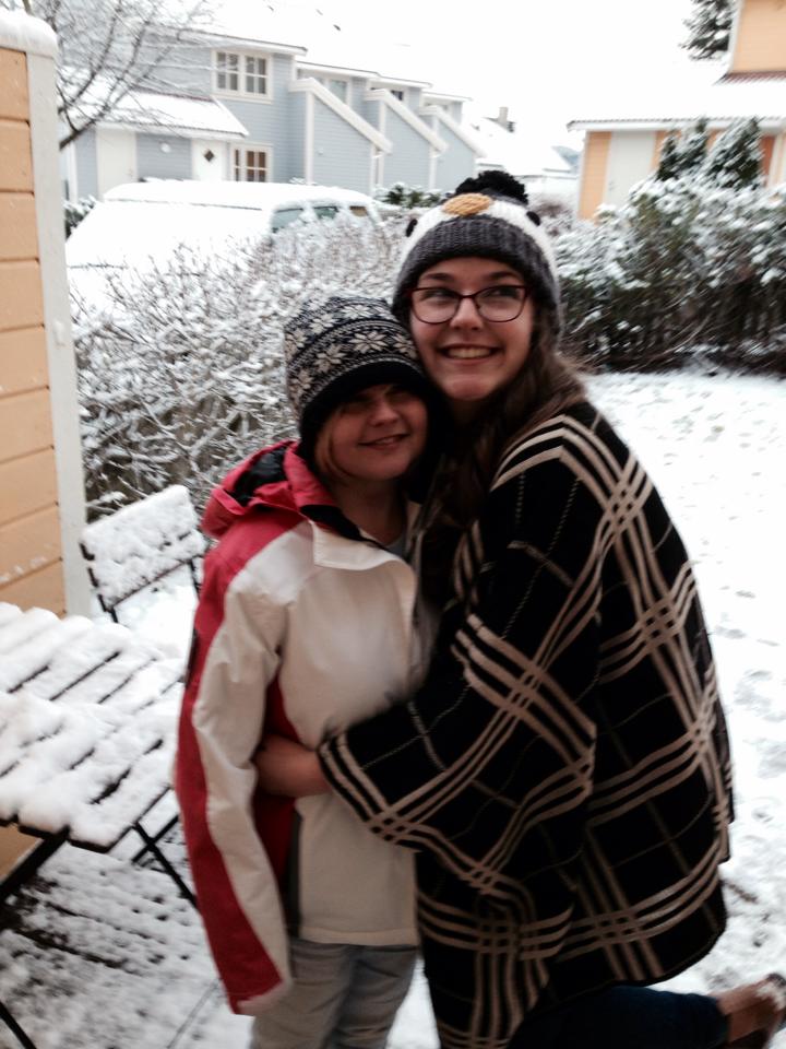 Sassy wearing a white and red superdry jacket and a bobble hat. Her sister is wearing a wollen poncho and also wearing a bobble hat. There is snow covering the garden in the background