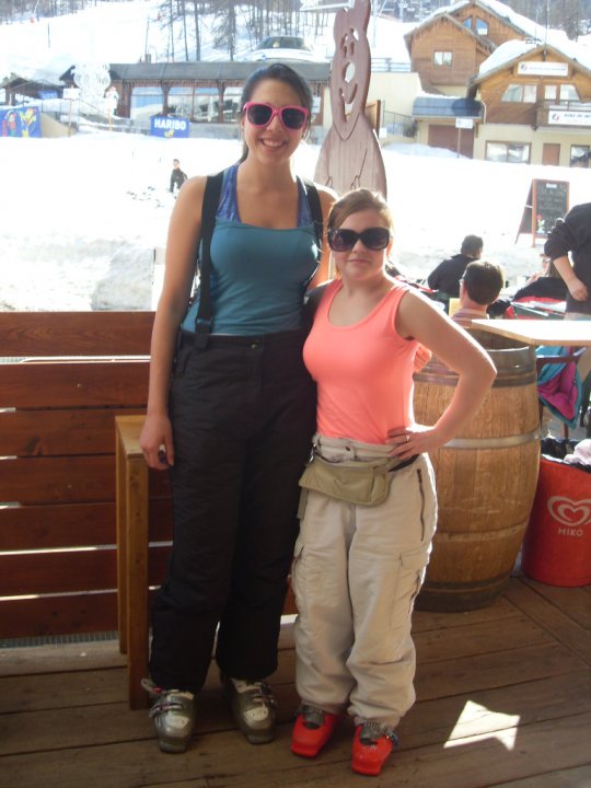 Sassy wearing a pink Tank Top, large sunglasses and red ski boots. Standing with Christine who is wearing a green tank Top and grey ski boots with black trousers