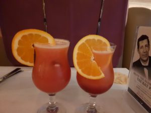 Two Red cocktails with big Orange slices garnishing the rim. These cocktails are Shimla Passion they contain Vodka, passoa, cranberry juice and orange juice