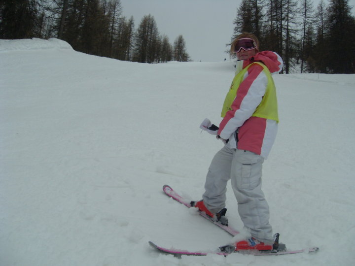 Sassy wearing white trousers, a pink and white jakcket and pink ski goggles. She is standing at the bottom of a ski slope.