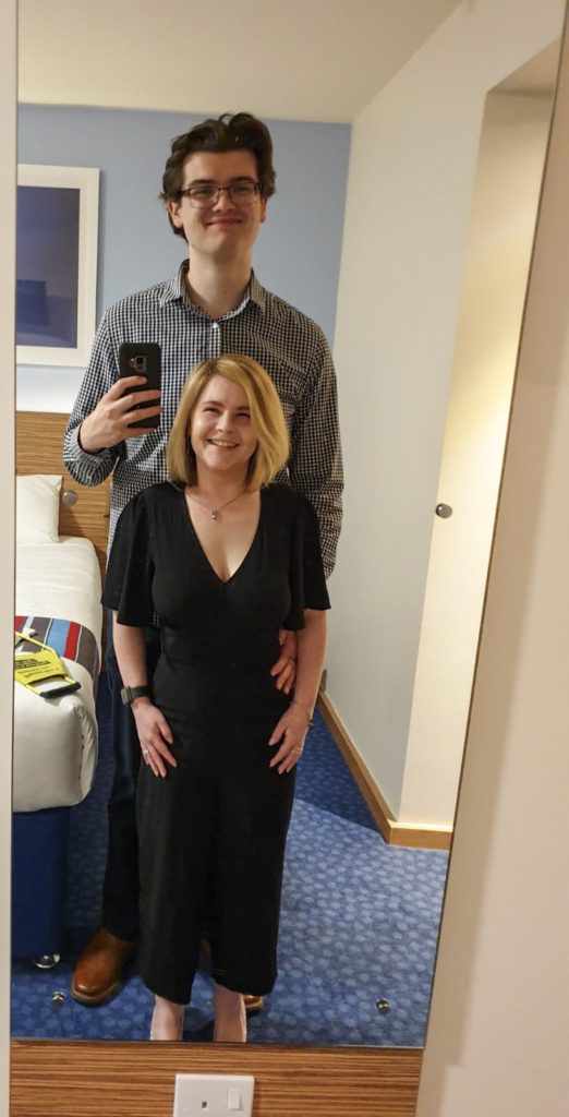 Sassy and Grant in front of a mirror in a hotel room, smiling. Grant is behind Sassy, holding the pone to take the photo. Sassy is wearing a long black dress, whilst Grant is wearing a black and white shirt, and black trousers.
