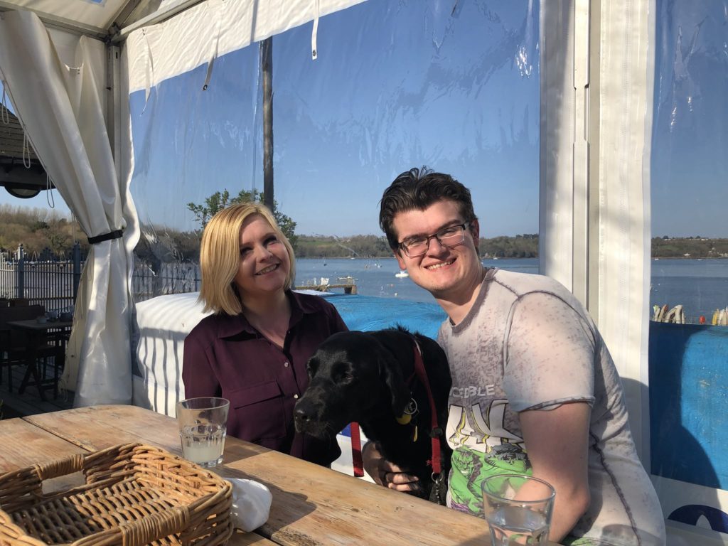 Sitting at a wooden bench style table, Sassy and Grant both smile towards the camera. In between them, with Grant's arm around her, Ida has her front paws up on the bench, with her nose straining towards the top of the table. In the background, through the clear plastic of the gazebo style covering, the blue water of the River Dart is visible, with small boats upon it.