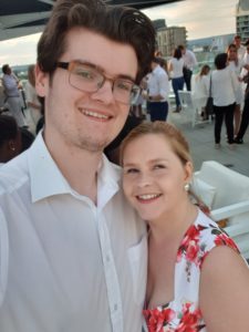 Sassy and Grabt are on a roof top, Grant is smiling towards the camera and so is Sassy. Grant is wearing a white shirt and Sassy is wearing a white dress with flowers.
