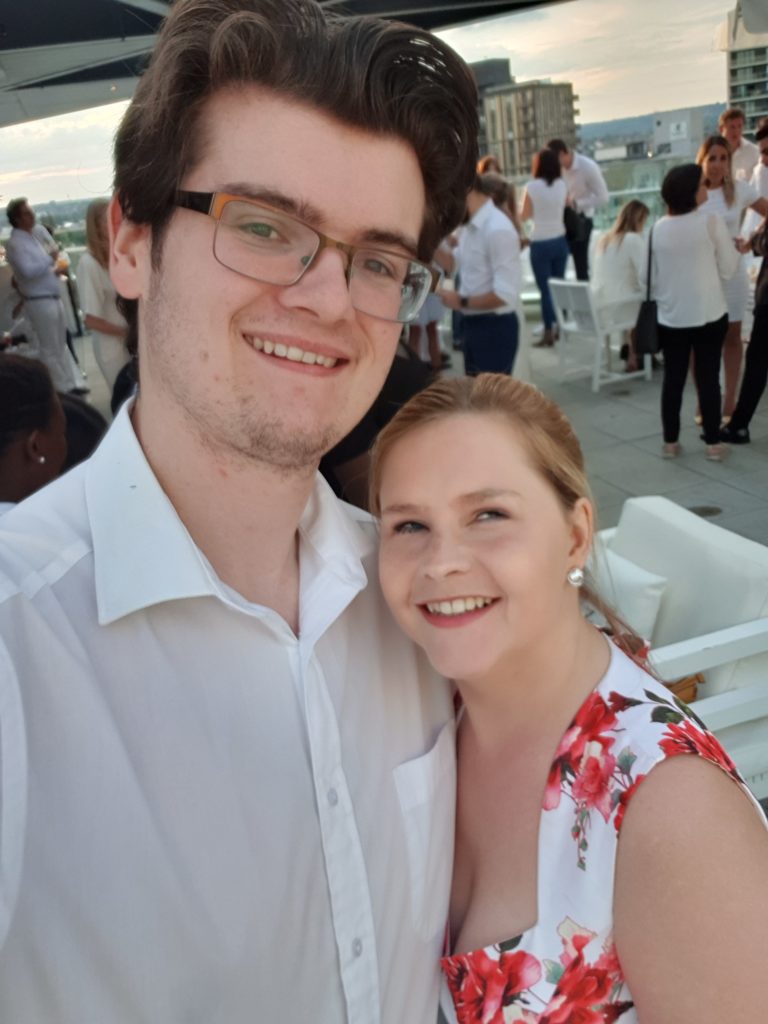 Sassy and Grant are on a roof top, Grant is smiling towards the camera and so is Sassy. Grant is wearing a white shirt and Sassy is wearing a white dress with flowers.