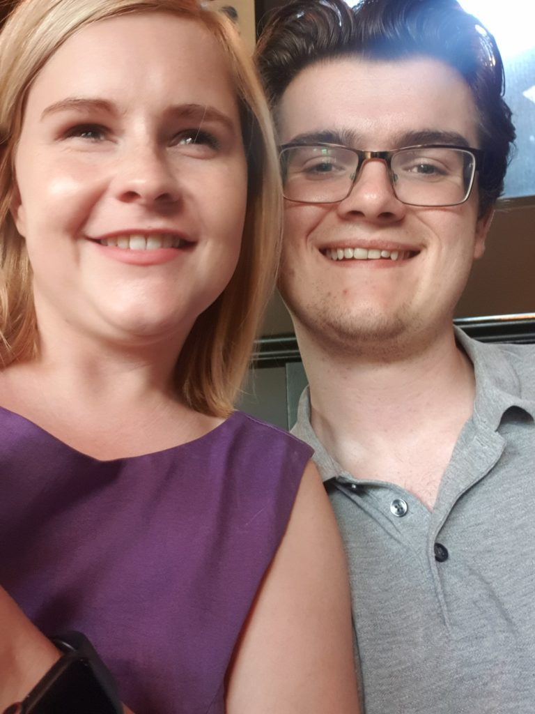 Sassy is wearing a purple dress and is smiling towards the camera, Grant is sitting next to Sassy and leaning in they are both smiling