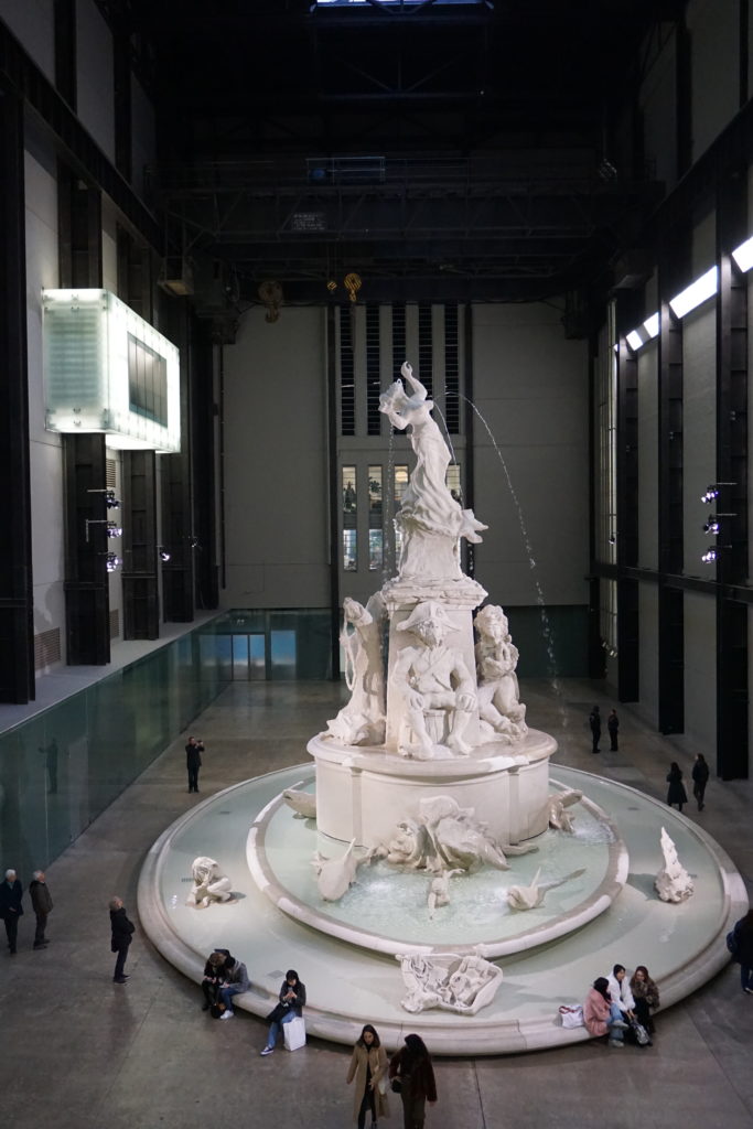 A Giant marble statue in the Tate Modern, there is water spraying out of a woman's mouth, they water is arching down into two pools below. There are marble sharks a boat and mermaids.