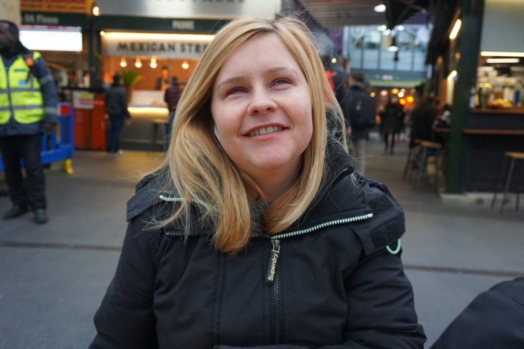 Sassy wearing a black coat, her blonde hair falls below her shoulders, she is smiling towards the camera. Market stalls are blurred in the background