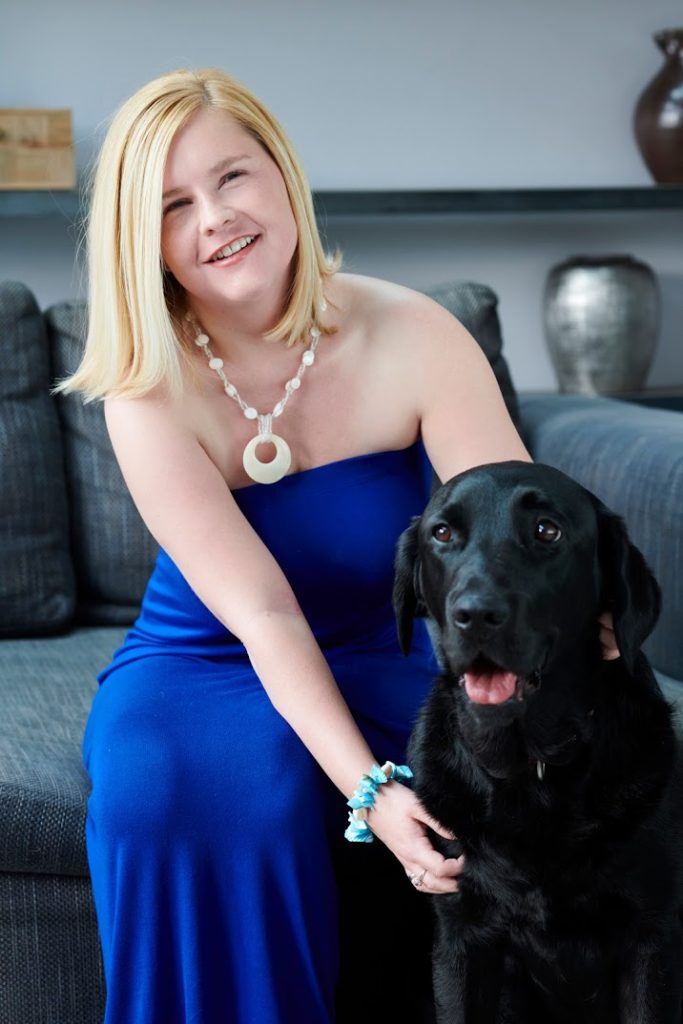 Sassy in a dark blue dress, she has long blond hair and is stroking her black lab Ida