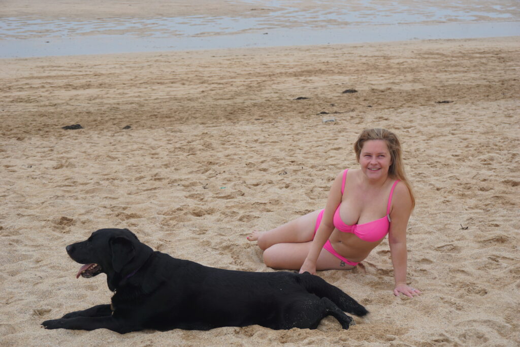 Sassy wearing a pink bikini lying on the beach with Ida, her black Lab lying down next to her.