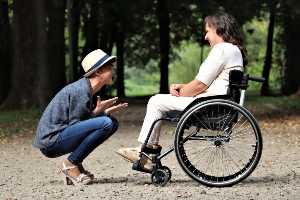 A lady who is in a wheelchair is on a woodland path, another lady is croughting down to talk to the lady in the wheelchair