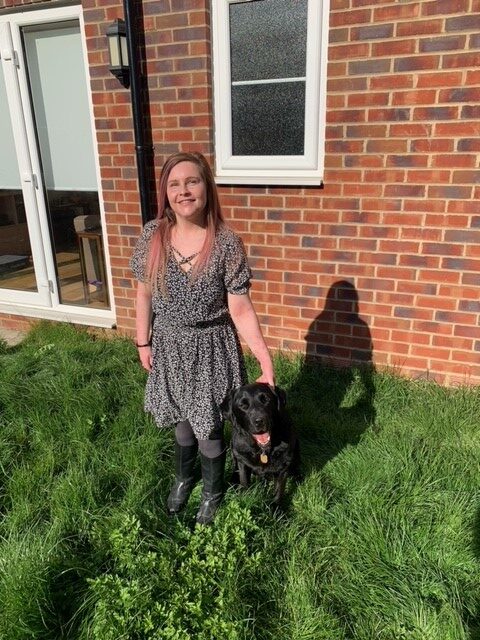 Sassy, A petite white woman with long purpley pink hair, wears a black and white floral dress with capped sleeves, with black tights and long black boots. Standing in the garden the Sun shines down upon her. Ida her black lab retriever cross sits on her left and Sassy’s hand rests on Ida’s head. Sassy smiles towards the camera their shadows cast on the wall behind them.