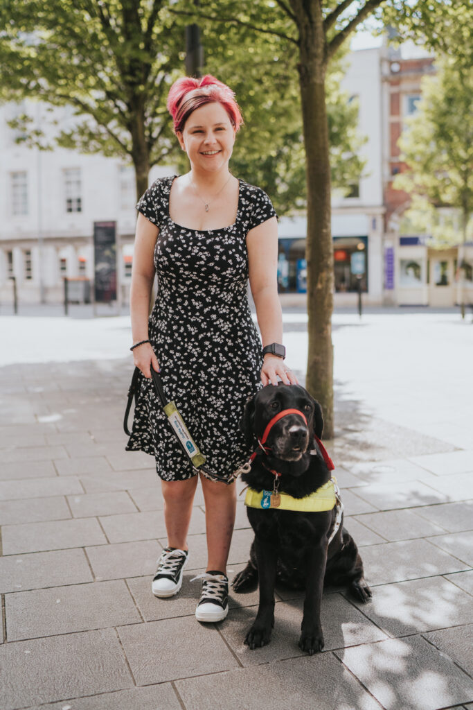 Sassy wearing a black dress with white flowers, stands next to Ida a Black Labrador retriever cross, she is wearing her guide dog harness and holty.There are two trees and a row of shops in the background.