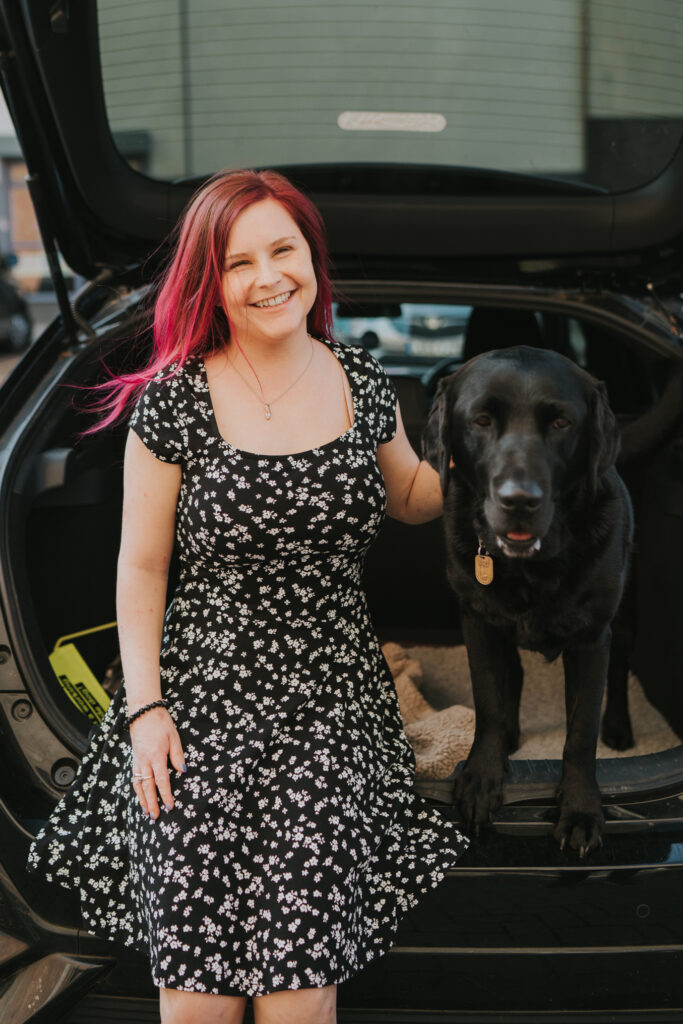 Sassy in her black and white floral dress stands with Ida who has her front paws on the lip of their car. Sassy is smiling towards the camera and Ida is look straight ahead.
