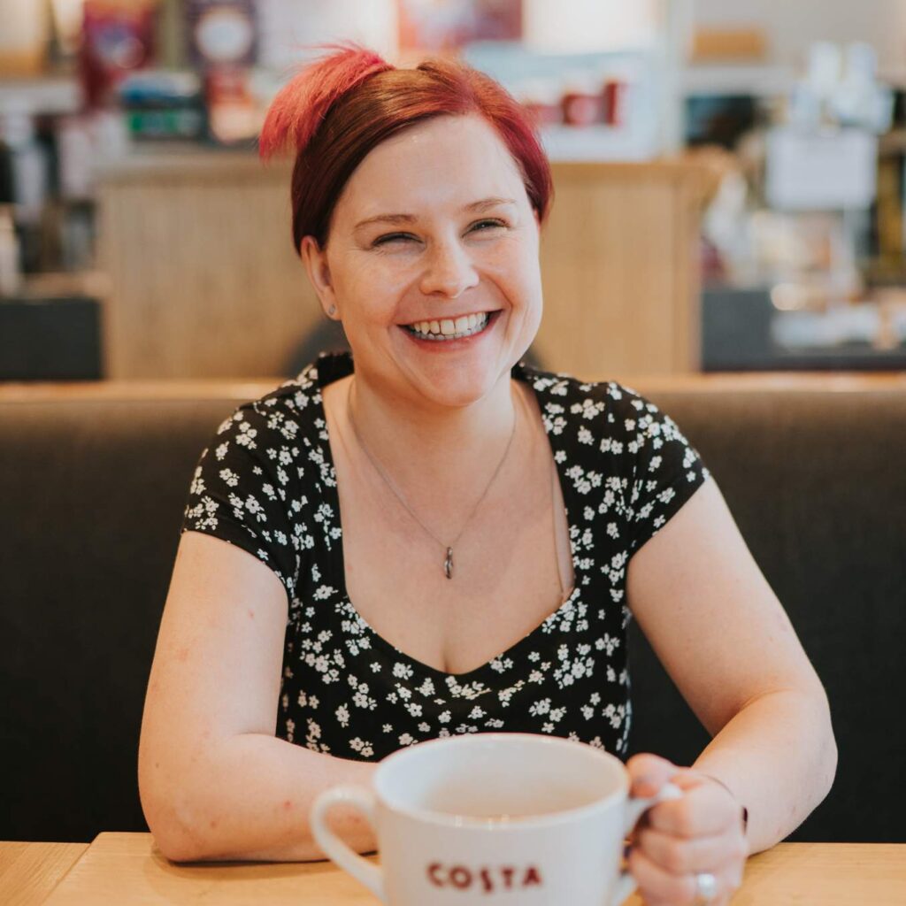 Sassy wearing a black dress with white flowers, sitting down in a coffee shop, holding a large mug of coffee, and smiling at the camera.