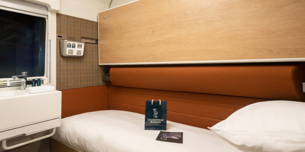 Caledonian sleeper train club room. You can see a single bed with a blue bag that says caledonian sleeper on top. In the bottom right corner you can see the pillow and in the top left corner is the window, where it's dark outside.