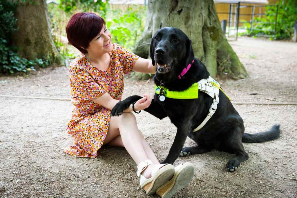 Sassy sitting on the ground, wearing a flowery dress and white sandals, holding Ida's paw. She is looking towards Ida, smiling. Ida is wearing a reflective harness and a pink collar.