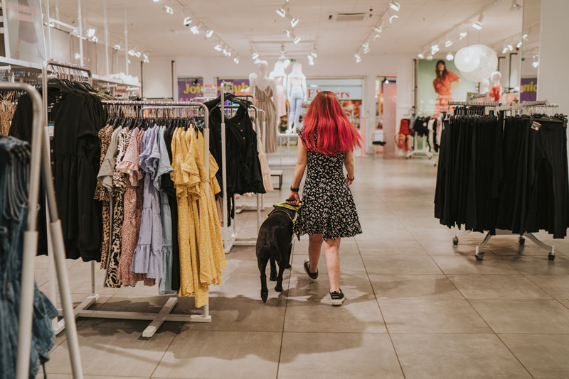 Sassy wearing a black dress with white dots, walking away from the camera in a clothes shop. She is guided by Ida the black guide dog, who is on her left. Sassy has long red hair and is with the back at the camera.