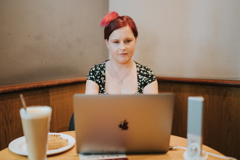 Sassy sitting down in a coffee shop, concentrating on the laptop in front of her. There is a large glass of latte and a plate with a sandwich on the table as well. Sassy is wearing a black dress with small white flowers and has her red hair up in a semi-bun.