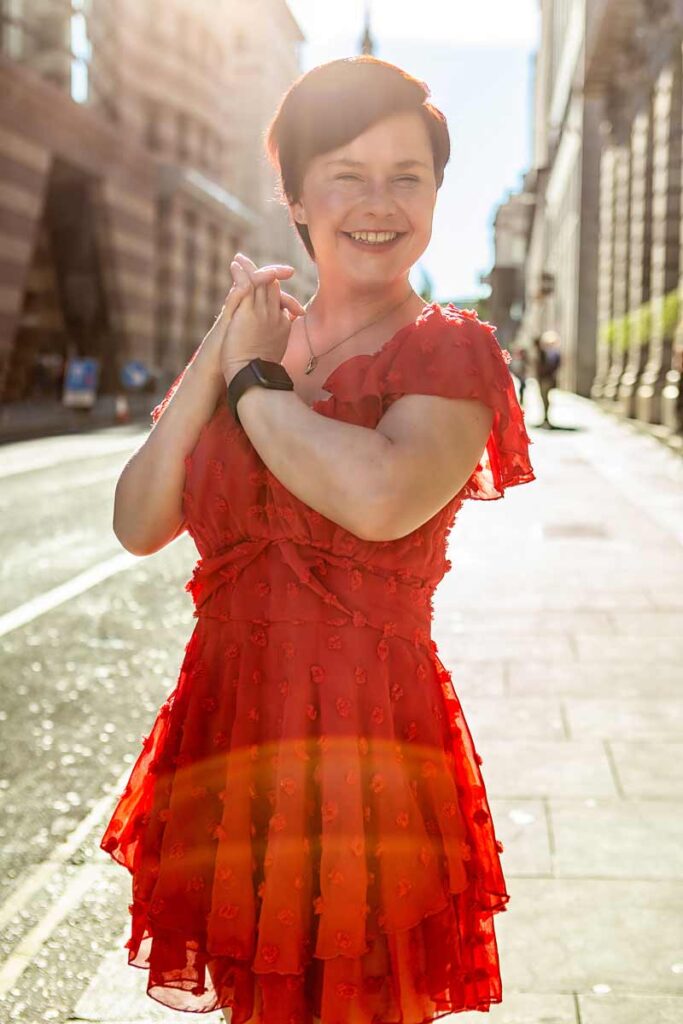 Sassy wearing a red dress and a smart watch on her left wrist, holding her hands together and smiling at the camera. She has short red hair, and the light around her is creating a warm halo effect.