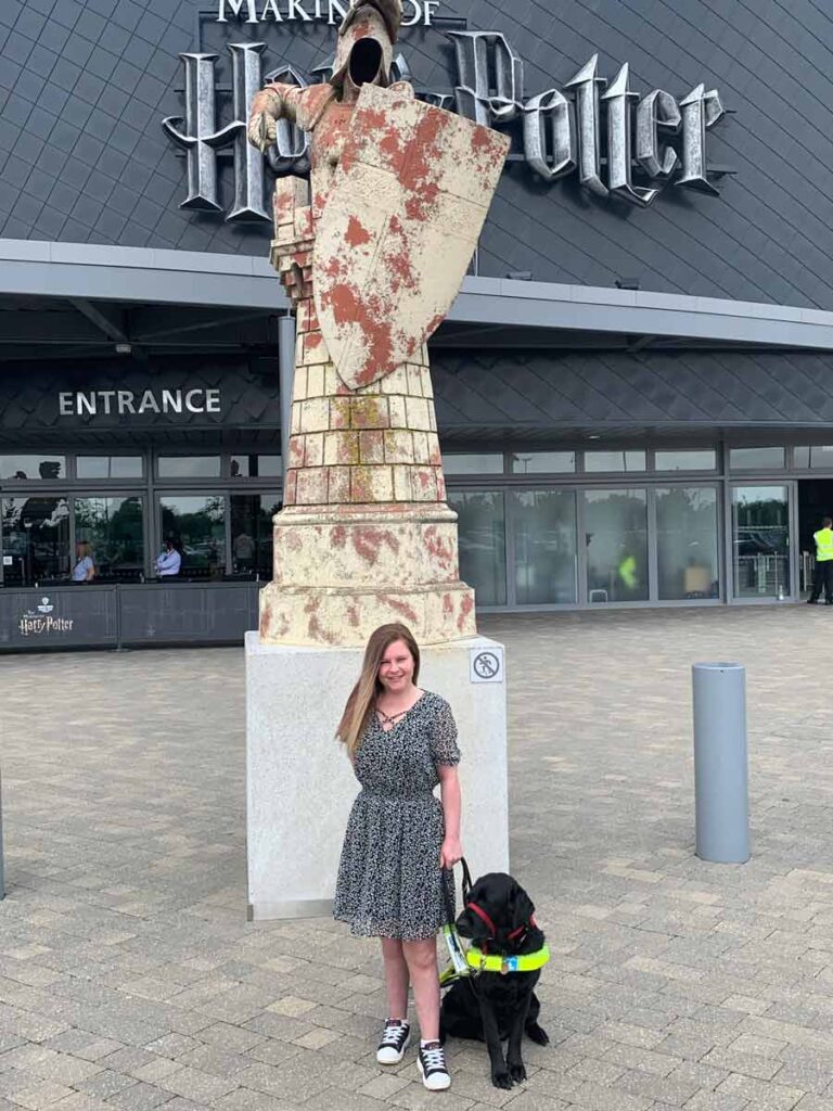 Sassy wearing a knee long black dress with small white flowers, smiling at the camera and holding Ida's leash. Ida is sitting by her side, on the right hand side. Behind Sassy there is the entrance to Harry Potter Studio.