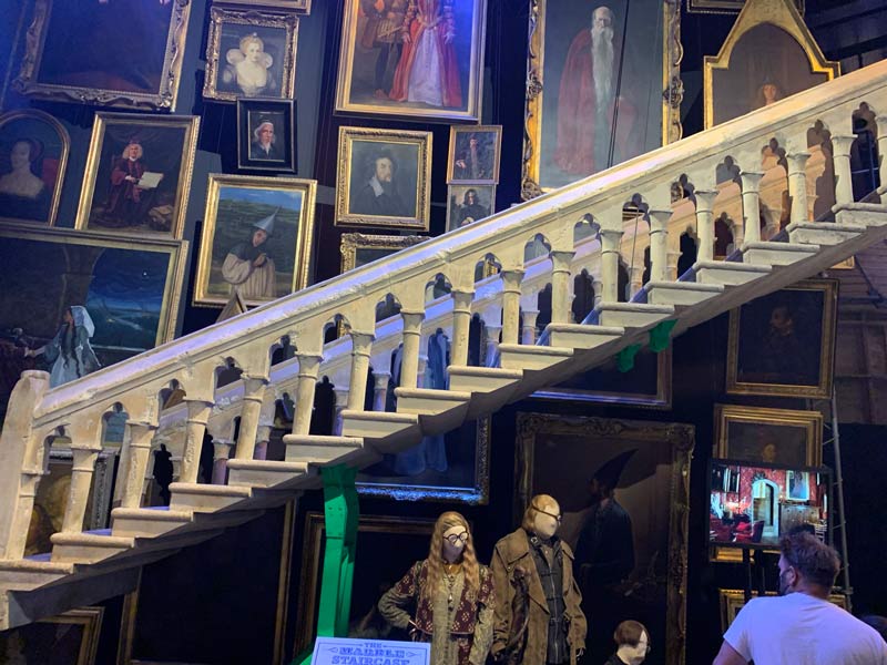 A staircase supported by small columns which look Roman. Above the staircase there are a lot of paintings of different wizards. Below, there is an exhibition with costumes from the movie.