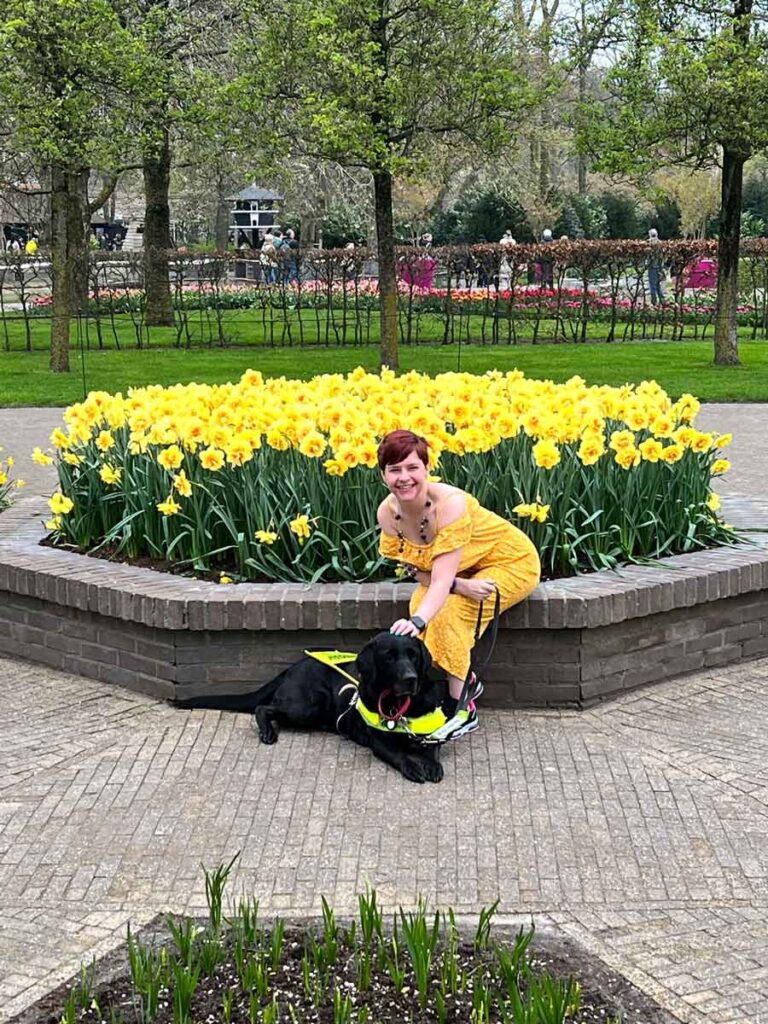 Sassy posing with Ida the black Labrador in front of a large bunch of yellow daffodils. She is wearing a yellow jumpsuit and is sitting down on the stone edge of the garden.