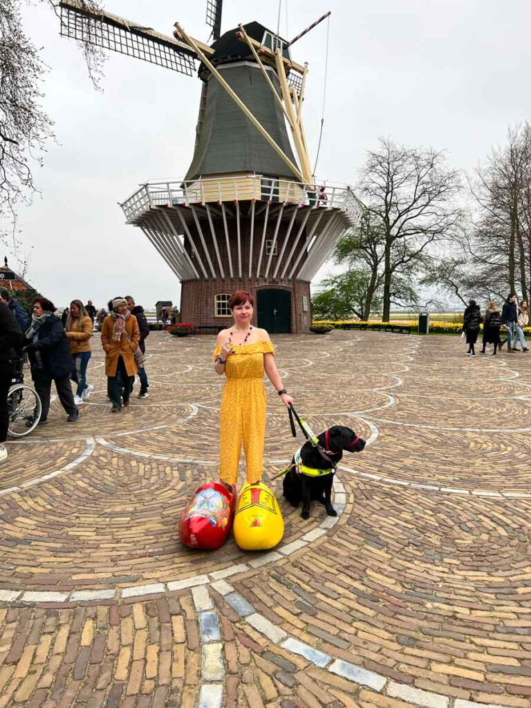 Sassy wearing a yellow jumpsuit and a black necklace, posing in front of a windmill. She has her legs inside oversized clogs, one red and one yellow. Ida the black Labrador is at her side.