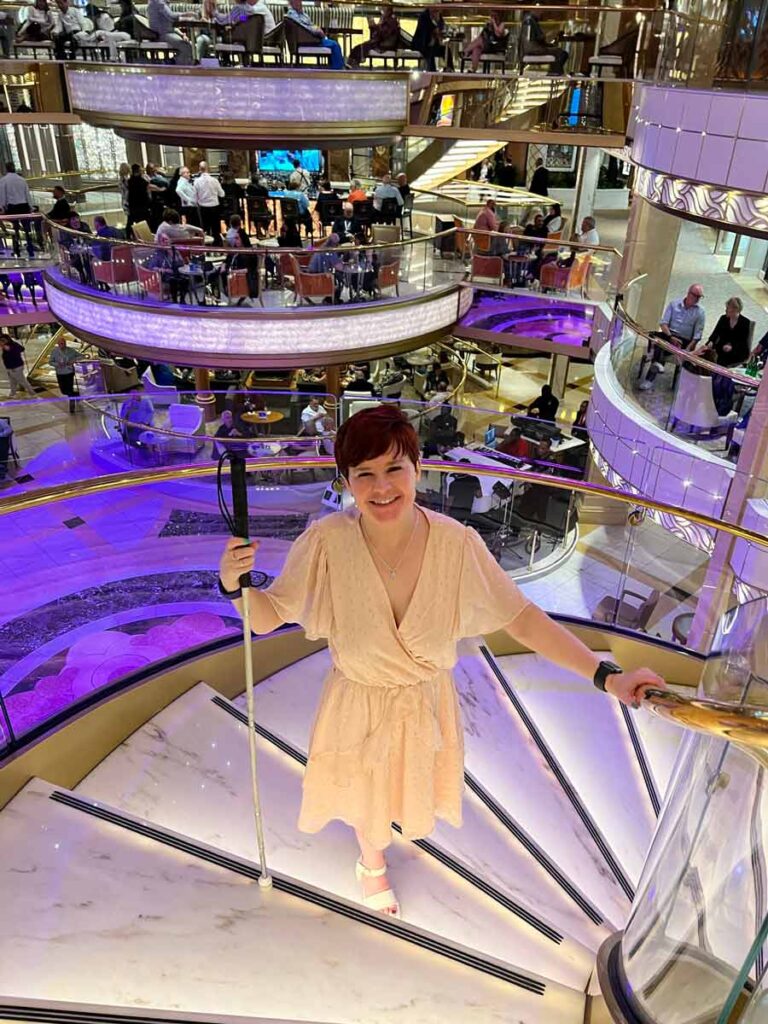 Sassy posing on the staircase of the ship. She is wearing a light coloured dress, white sandals, and is holding her cane in the left hand. She is smiling at the camera. Behind, you can see a large area with balconies with chairs and tables, all illuminated in a purple light.