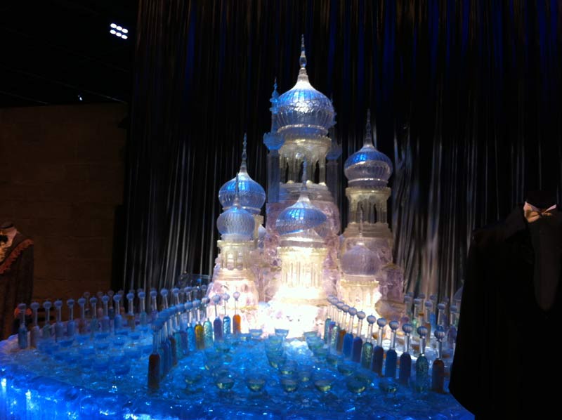 A palace that looks like it is made from glass or ice. It is illuminated in blue lights. At the bottom there are many glasses filled with different coloured liquids.