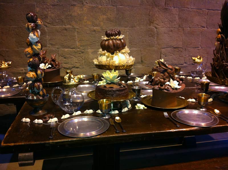 A wooden table filled with different cakes and desserts. Along the plates there are small white mice arranged in a row, like they are following each other. The cakes have different shapes: one is a rabbit on top of a hat, another one is made from pumpkins on top of each other.