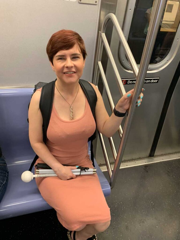 Sassy wearing a long orange dress and a black backpack, sitting down on the train in New York. She has her nails painted light blue. She is smiling at the camera, whilst holding in her lap the folded white cane.