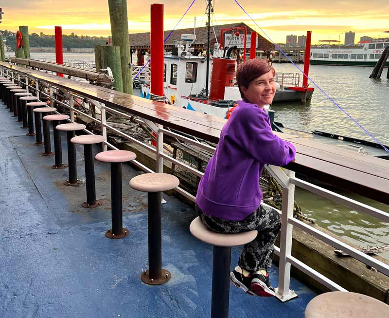 Sassy sitting on a bar stool, on an outdoors terrace, half turned towards the camera, smiling. She is wearing black and white trousers and a purple jumper. Behind her, there is a harbour with a small boat anchored. It is sunset time and the sky is orange and yellow.