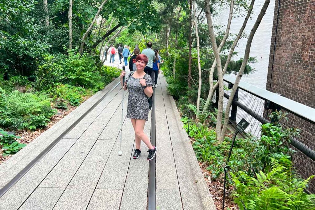 Sassy wearing a black short dress and black trainers with pink and white elements. She is wearing a black backpack and is holding her white cane whilst posing at the camera. She is inside a very green park