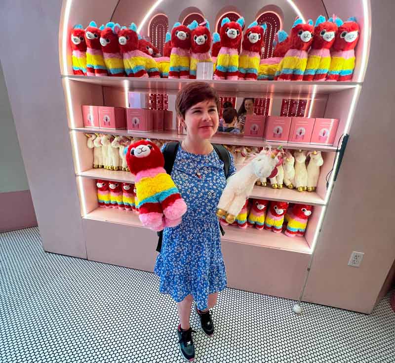 Sassy in a store selling colourful alpacas. There is an entire shelf with toy alpacas and toy unicorns behind her. She is wearing a blue dress with small white flowers, and is holding one alpaca in one hand, and an unicorn with golden hoofs in the other.