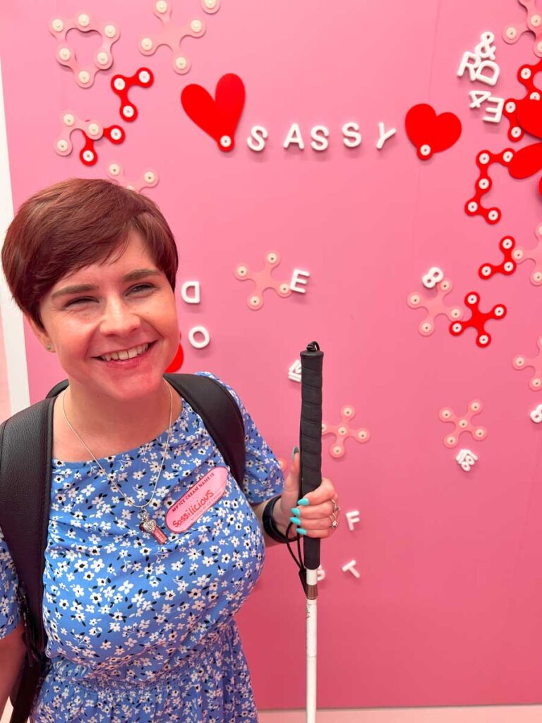 Sassy is wearing a blue dress with tiny white flowers, and has light blue painted nails. She is holding the white cane with her left hand. She is smiling at the camera. Behind her, there is a pink magnetic board, with the name "Sassy" spelled out in white letters, and two hearts on each side of it.