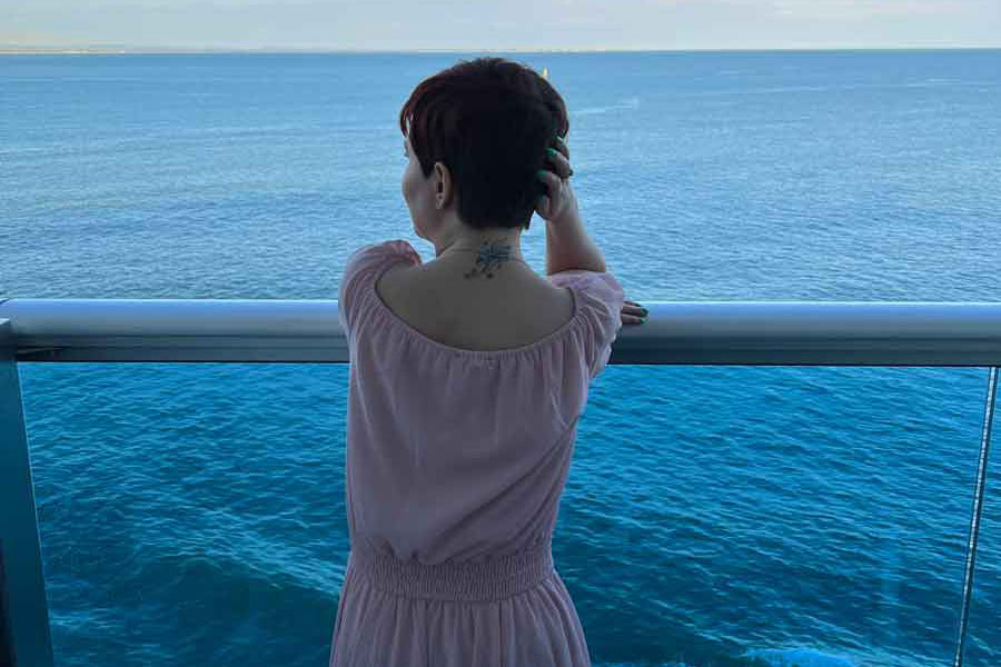A photo of Sassy wearing a pink long dress, looking towards the horizon from her balcony, at dusk. She is facing the sea and is with her back at the camera.