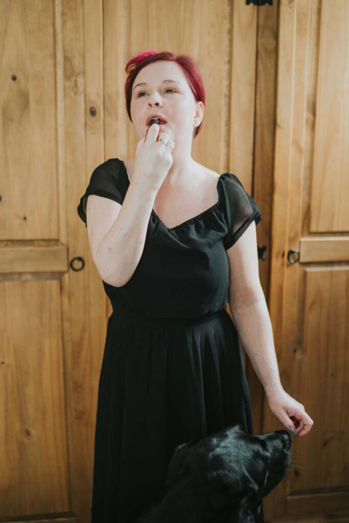 Sassy wearing a black dress with very short sleeves, applying lipstick to her lips. Her hair is red. You can just see Ida's head at the bottom of the photo, reaching for Sassy's arm.