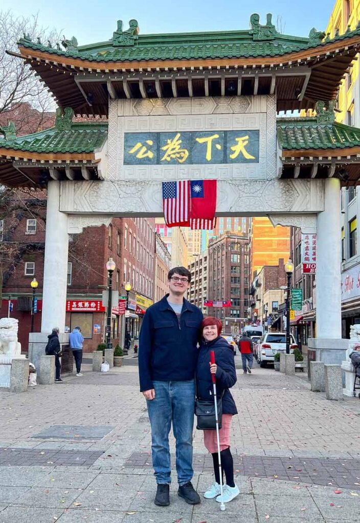 Grant and Sassy standing together in front of the gate of Chinatown, smiling to the camera. Grant is wearing blue jeans and a black jacket, whilst Sassy is wearing a pink dress and a black jacket on top. She is also holding a white cane. The roof of the Chinese gate is green.