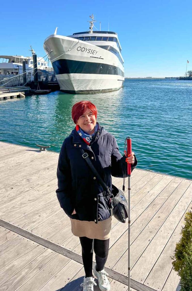 Sassy smiling at the camera, on a pier. She is wearing a cream skirt, black leggings, white trainers, a black jacket, blue and red scarf, and a black bag. She is holding her white cane, which has a red handle. Behind her, there is a large yacht named Odyssey anchored by another pier.