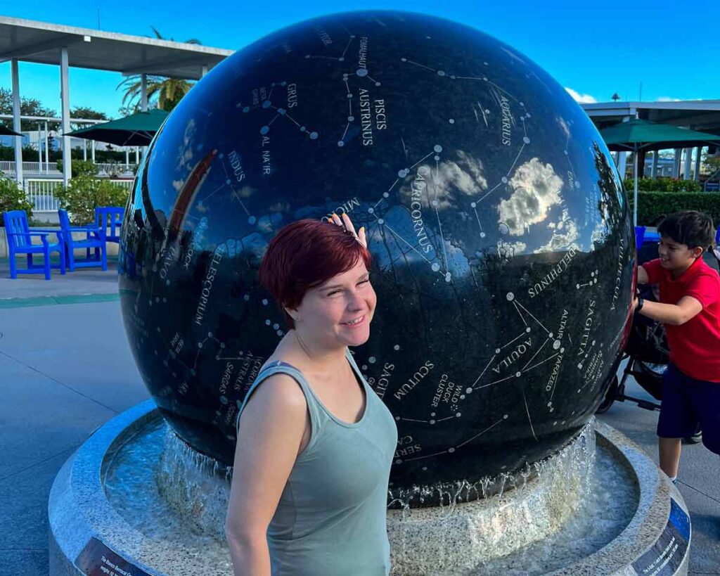 Sassy wearing a blue tank top touching a giant black ball with constellations on it. She is smiling.