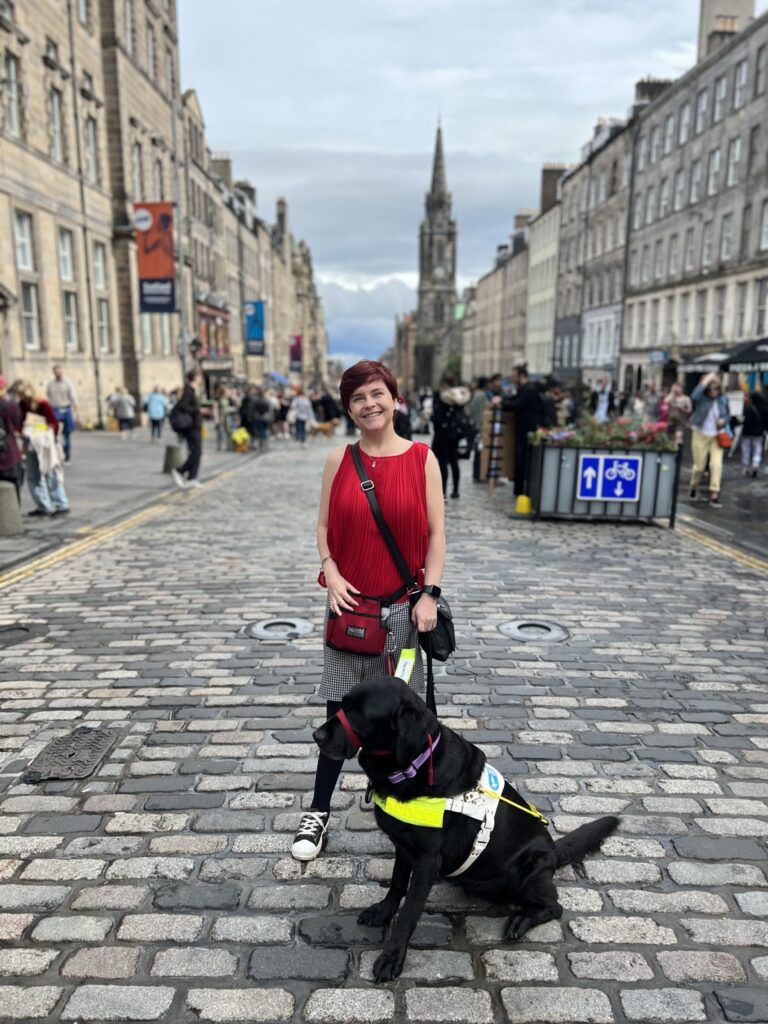Sassy in a red top, black checkered skirt and black tights with Ida a black Labrador in her guide dog harness stand on Edinburgh's Royal mile during the fringe festival.