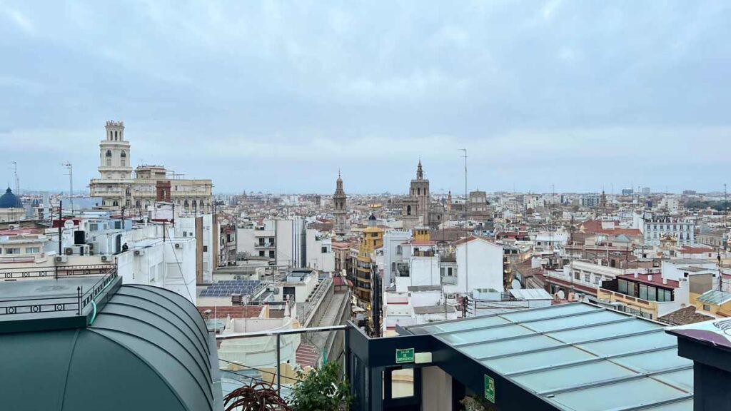 An image showing the city of Valencia from a height. You can see very different rooftops, showing the vast architecture of the city. There are medieval church towers in between modern building rooftops, some covered with metal, some having solar panels on top, and some being the typical Spanish terracotta red.