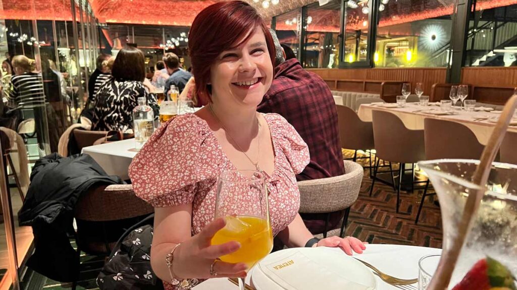 Sassy wearing a pink dress with white flowers, smiling at the camera from a restaurant. She has short red hair. She is holding a large glass with a yellow cocktail in one hand. Behind her there are tables with other diners.
