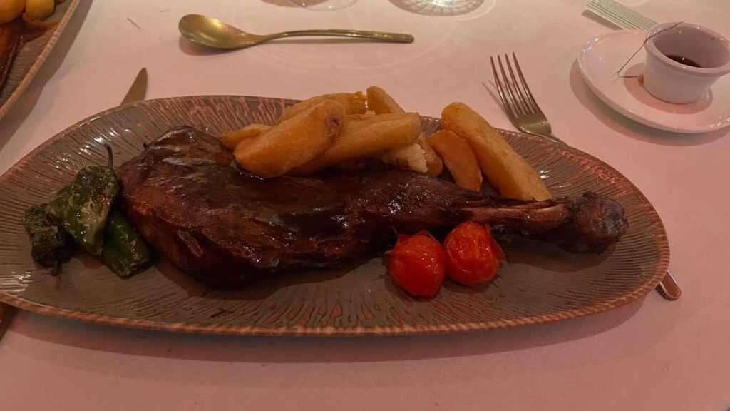 A large piece of meat on the bone with chunky chips, roast cherry tomatoes and pimentos del padron, on an oval dark green plate.